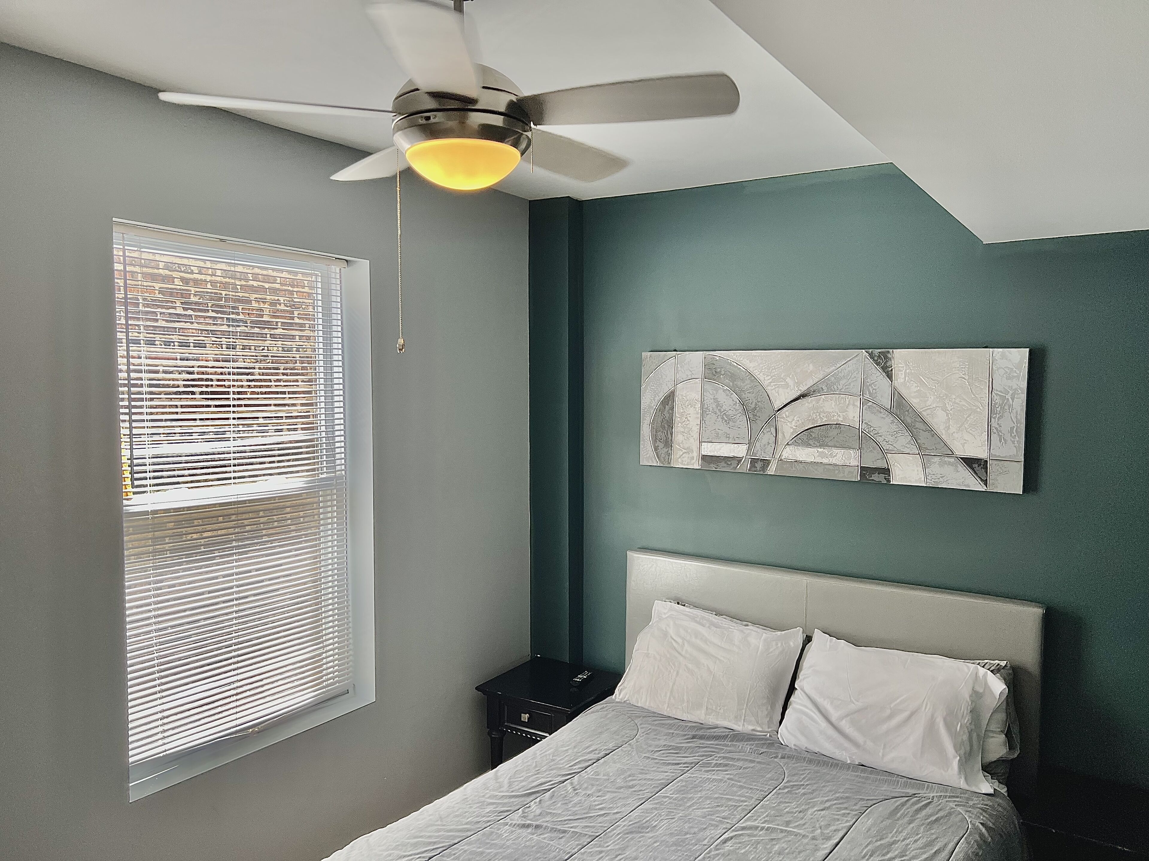 Quarto com cama, ventilador de teto, janela com persianas e uma obra de arte abstrata na parede.