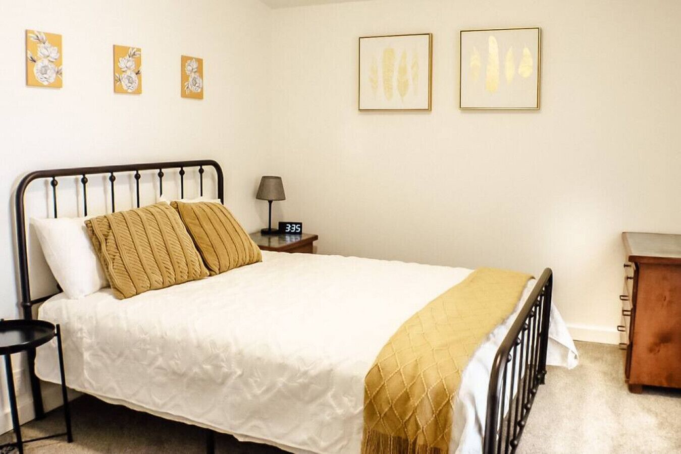 Ein ordentlich bezogenes Bett mit weißer Decke und zwei beigen Kissen, ein Nachttisch mit Lampe sowie zwei gerahmte Kunstwerke an der Wand.