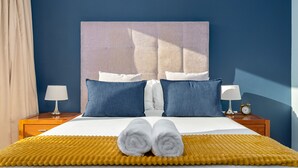 Una cama bien hecha con sábanas blancas, dos almohadas azules y un par de toallas blancas enrolladas sobre una manta amarilla con textura.