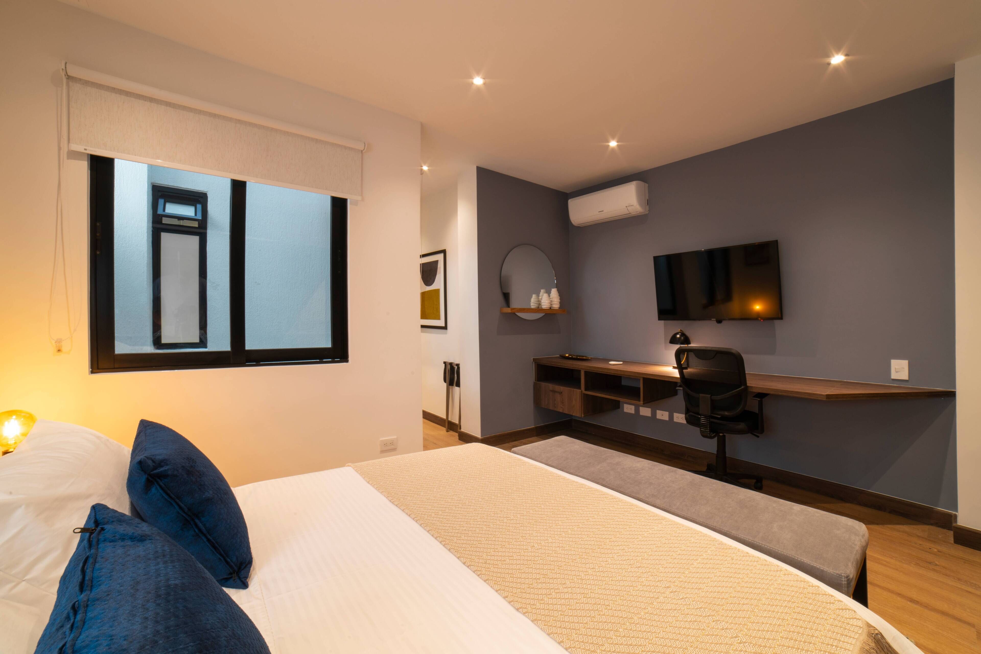 A modern hotel room with a large bed, a desk with a chair, a TV, and an air conditioning unit.