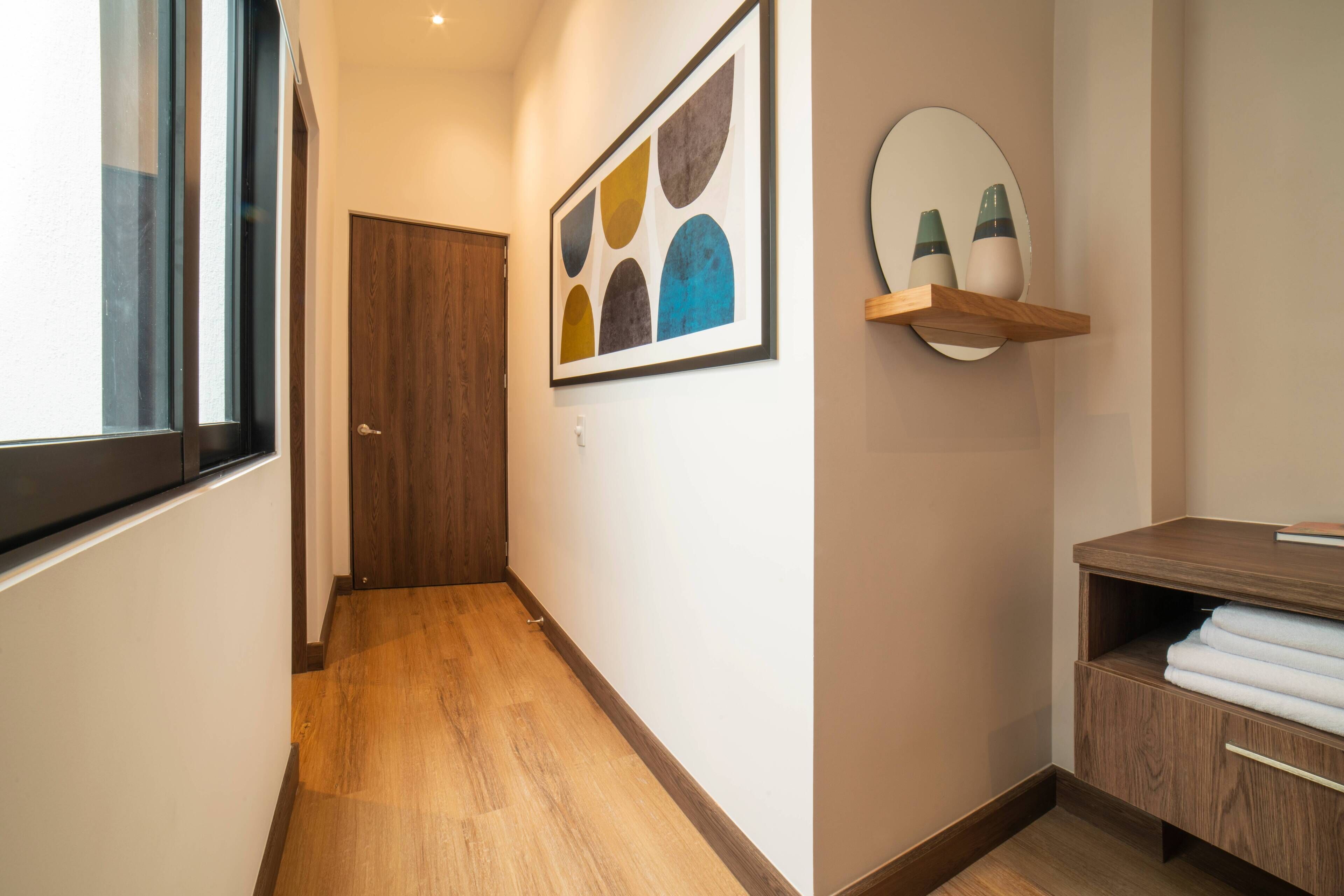 A hallway with a wooden floor, a door, a framed abstract painting, a mirror, and a wooden nightstand with towels.
