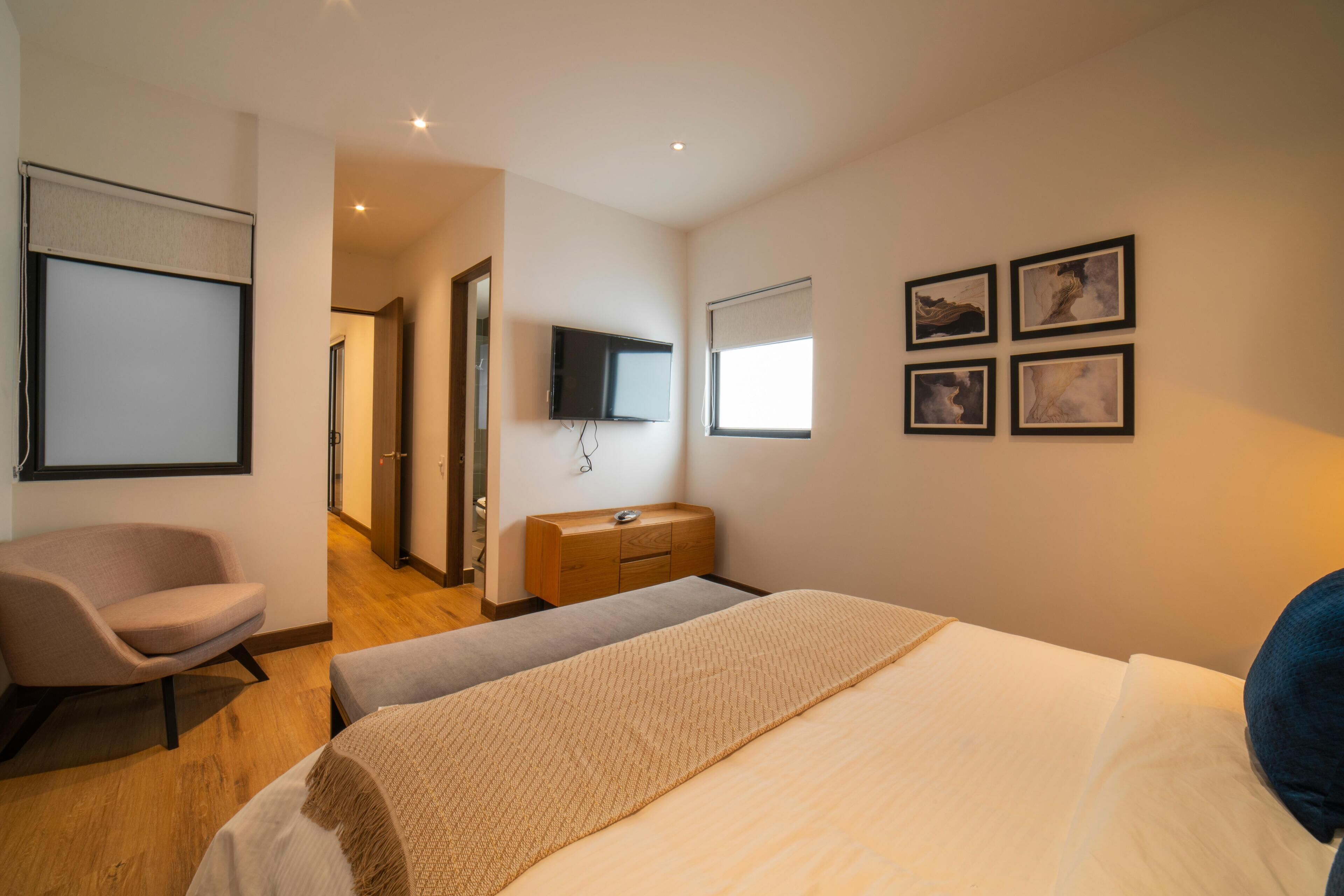 A hotel room with a bed, a chair, a dresser, a television, and framed pictures on the wall.