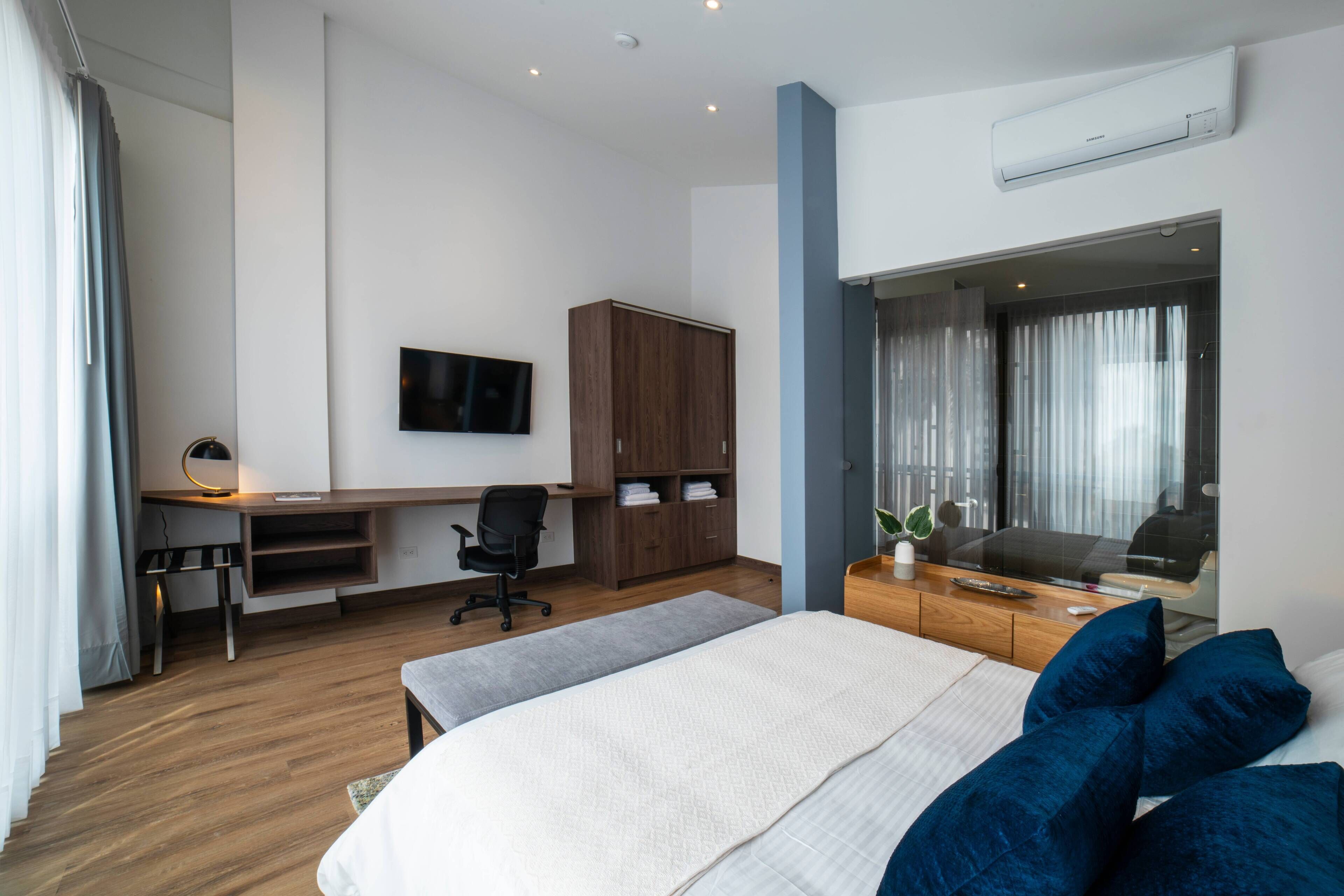 Ein modernes Hotelzimmer mit Bett, Schreibtisch, Stuhl, Kleiderschrank, Fernseher und einem großen Fenster mit Vorhängen.