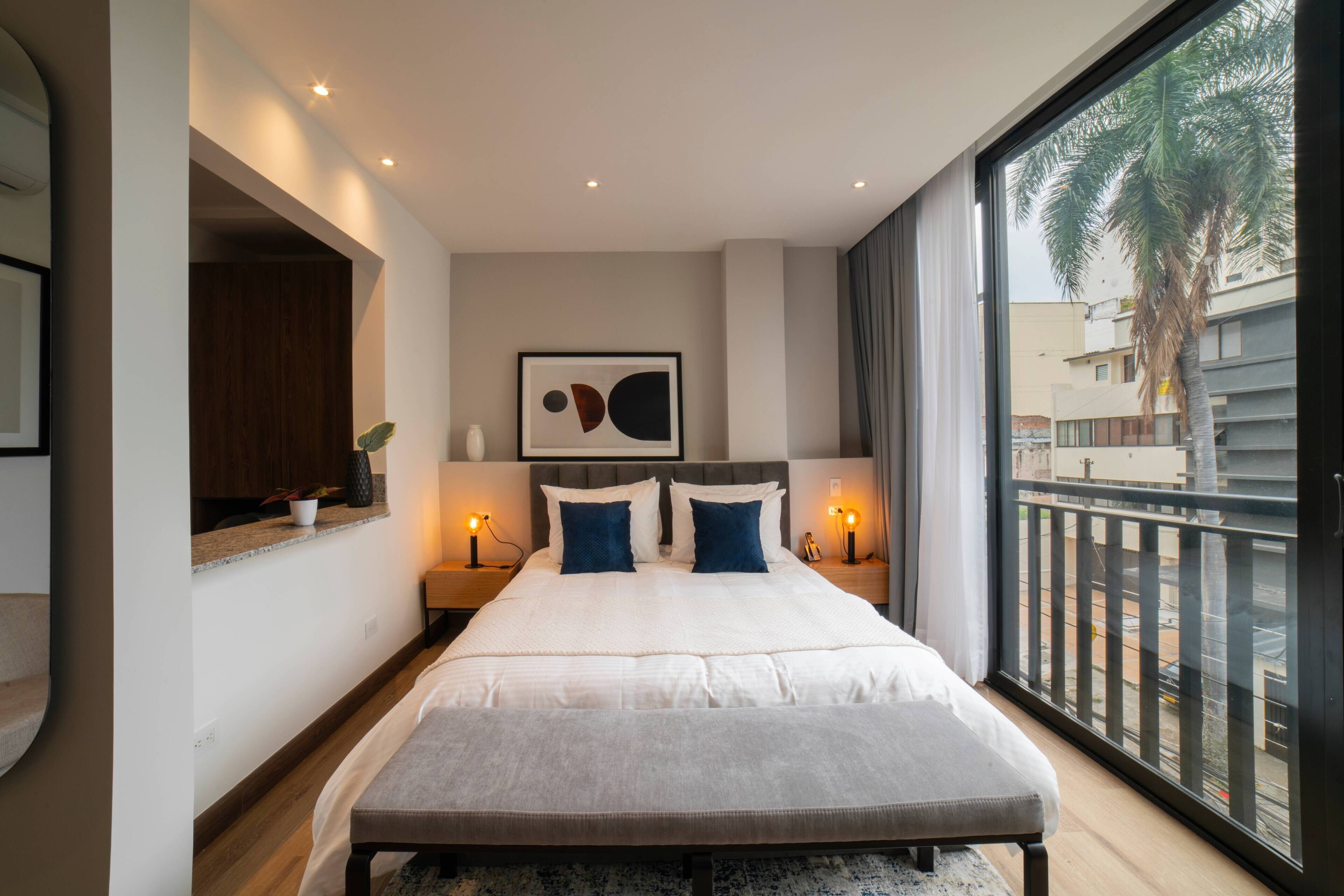 A modern bedroom with a large bed, bedside tables with lamps, a wall-mounted artwork, and a balcony with a view of palm trees and buildings.