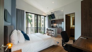 A modern hotel room with a large bed, a desk, a chair, a TV, and a kitchen area.