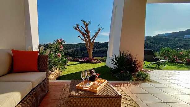 Uma área de estar moderna ao ar livre, com um sofá, bandeja com copos e comida, e vista para um jardim paisagístico e colinas.