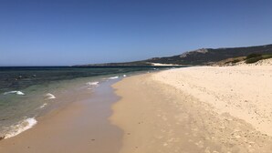 Uma praia com águas azul-claras, areal arenoso e colinas rochosas ao fundo.