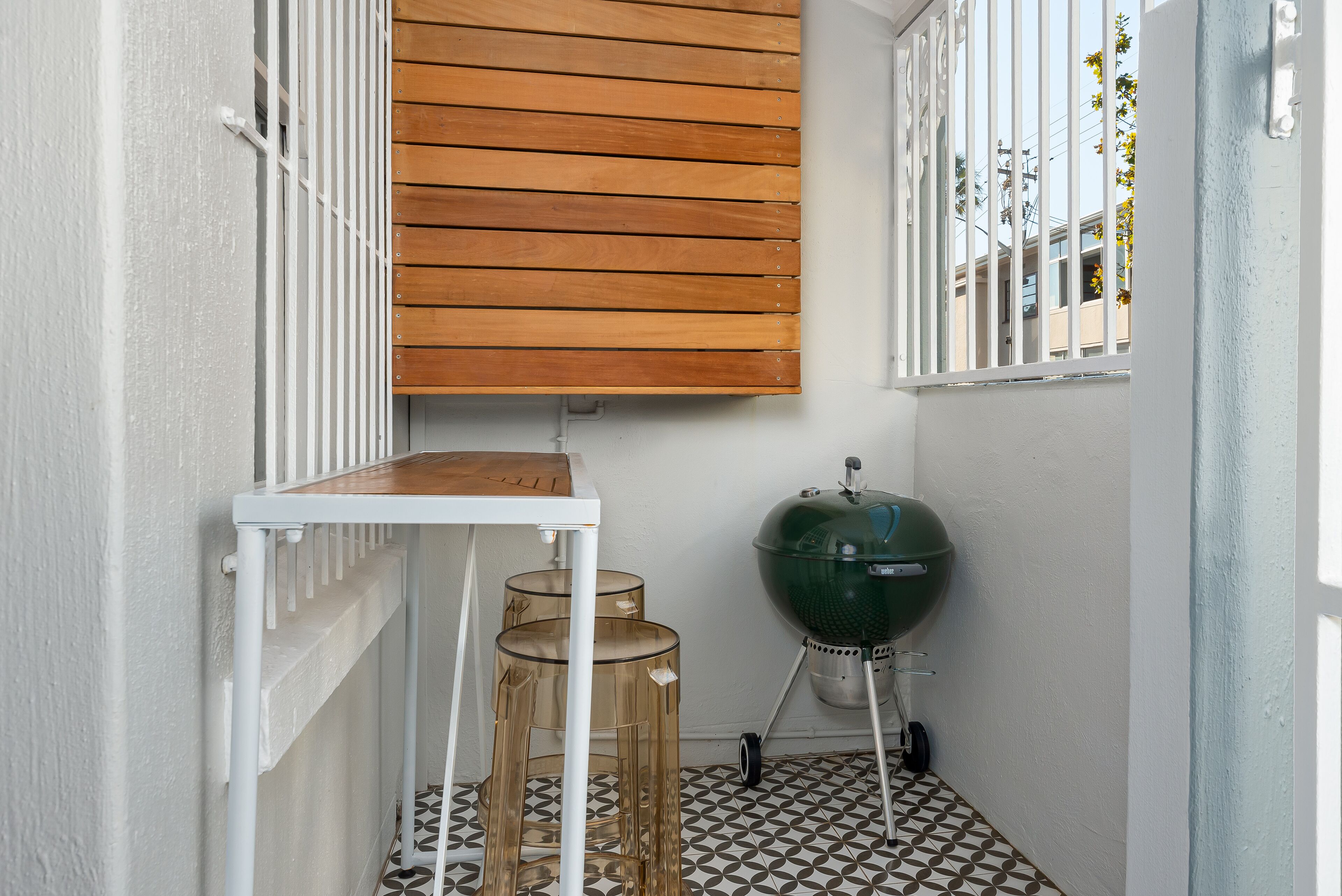 A small, enclosed outdoor space with a wooden slatted ceiling, a white table, two transparent stools, and a green barbecue grill.