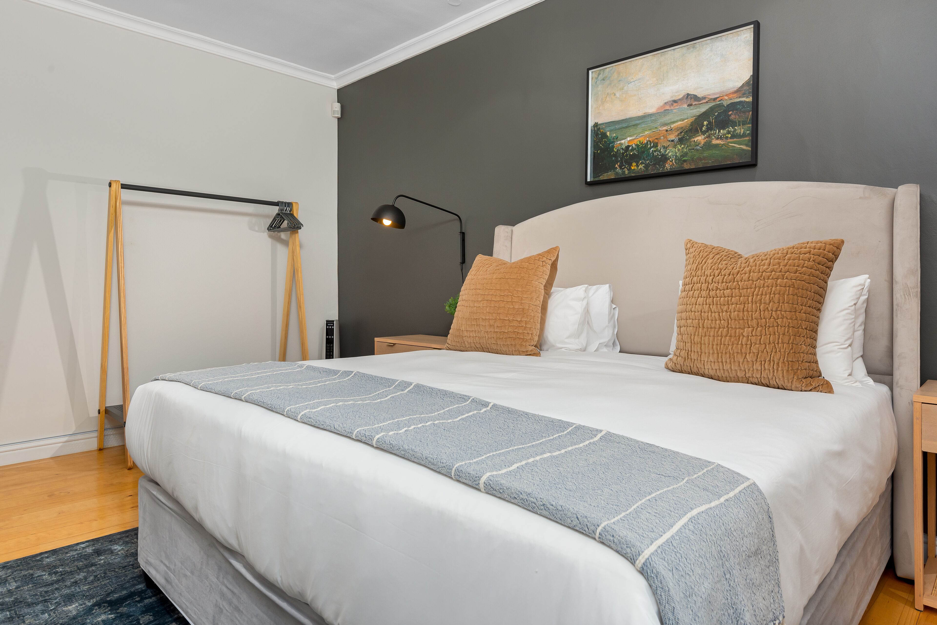 Cama bem arrumada com manta cinza e branca listrada, cabeceira com dois travesseiros, mesa de cabeceira com abajur e um quadro na parede.