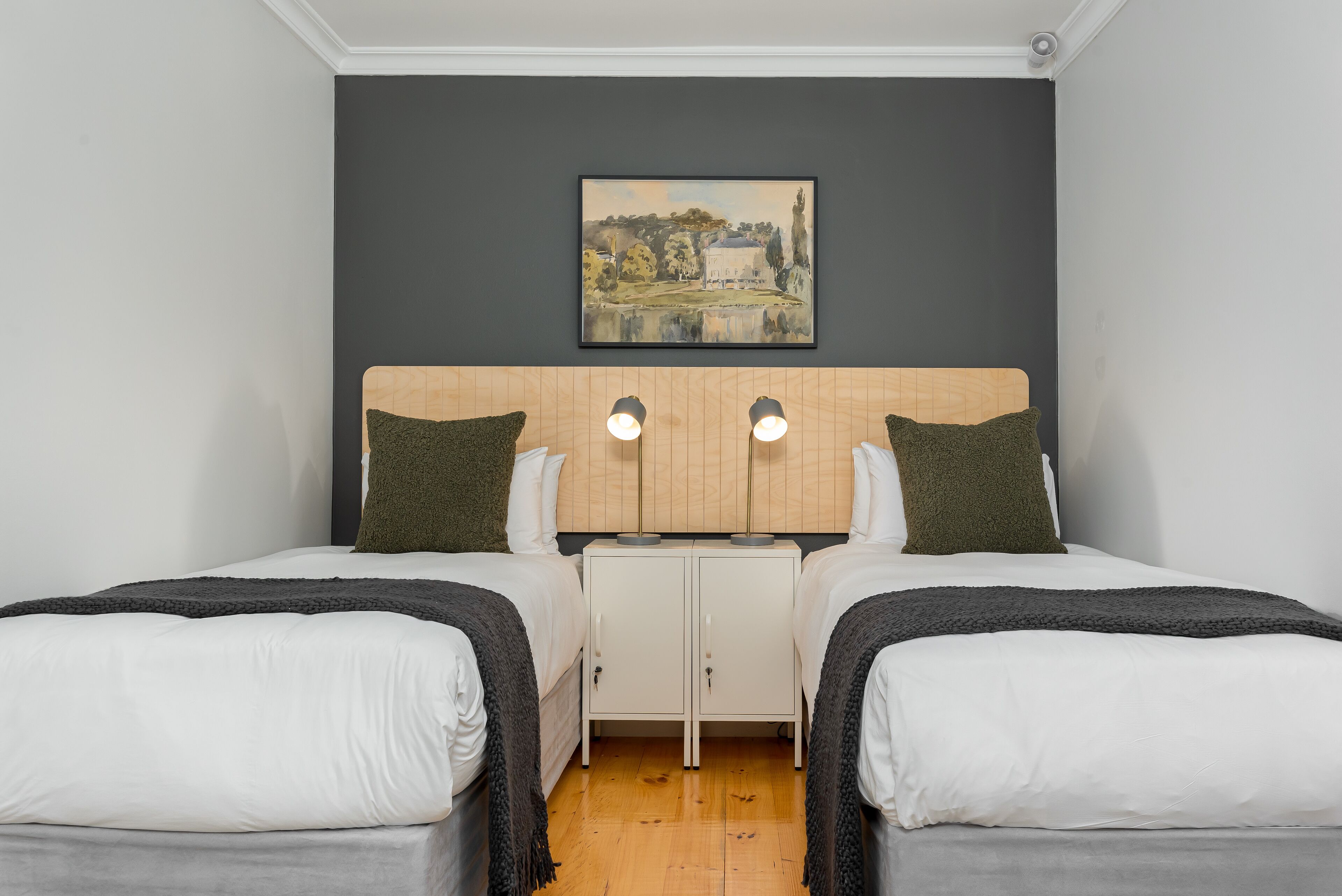 A twin room with two beds, a headboard with built-in cabinets, a painting on the wall, and a wooden floor.