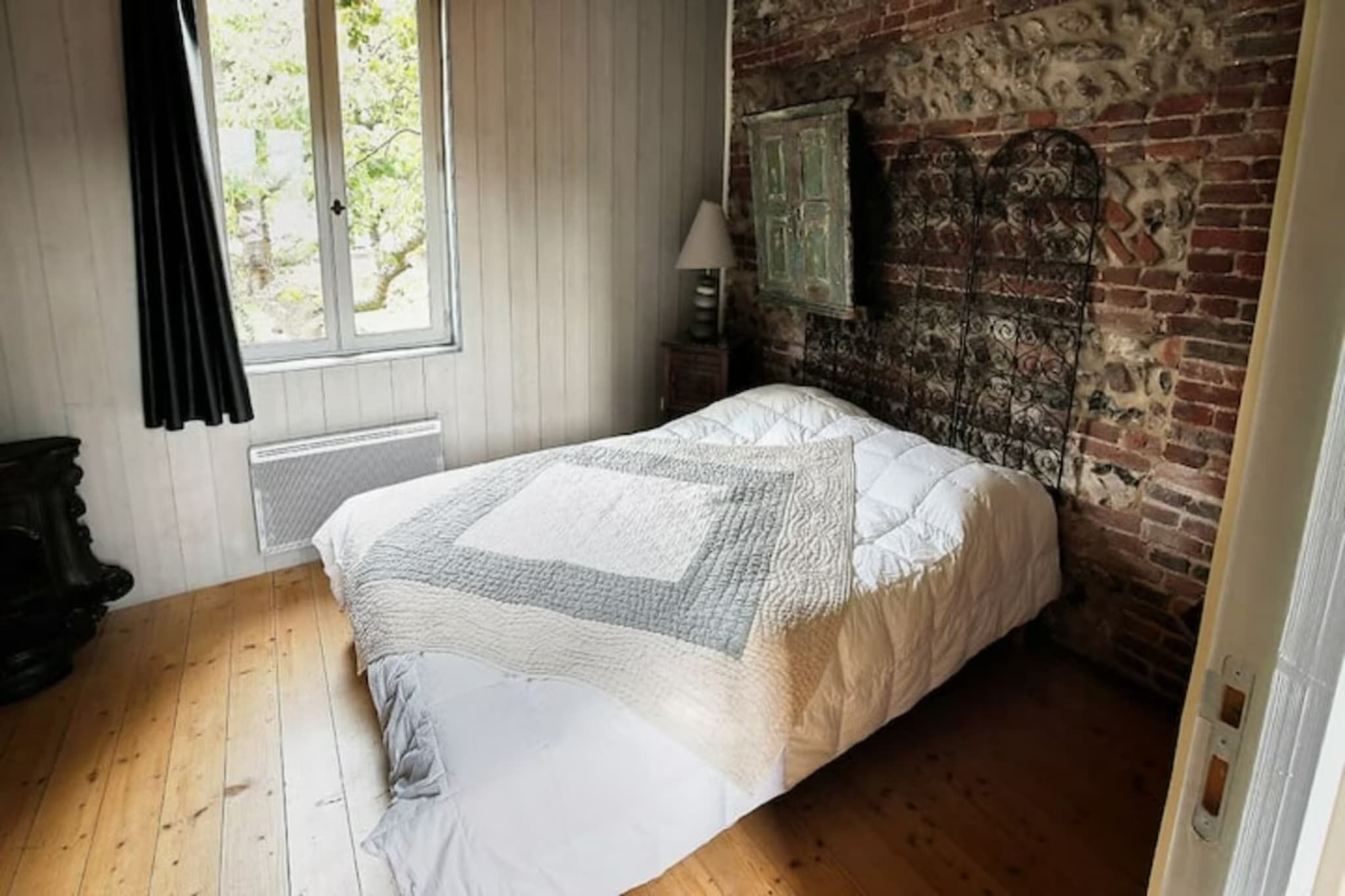 Un dormitorio con piso de madera, una cama con colcha, una ventana con cortinas, un radiador y una pared de ladrillo con cabecera de metal decorativa.