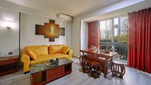 Una sala de estar con un sofá amarillo, una mesa de centro de madera, una mesa de comedor con sillas, un ventanal con cortinas rojas y una obra de arte colgada en la pared.