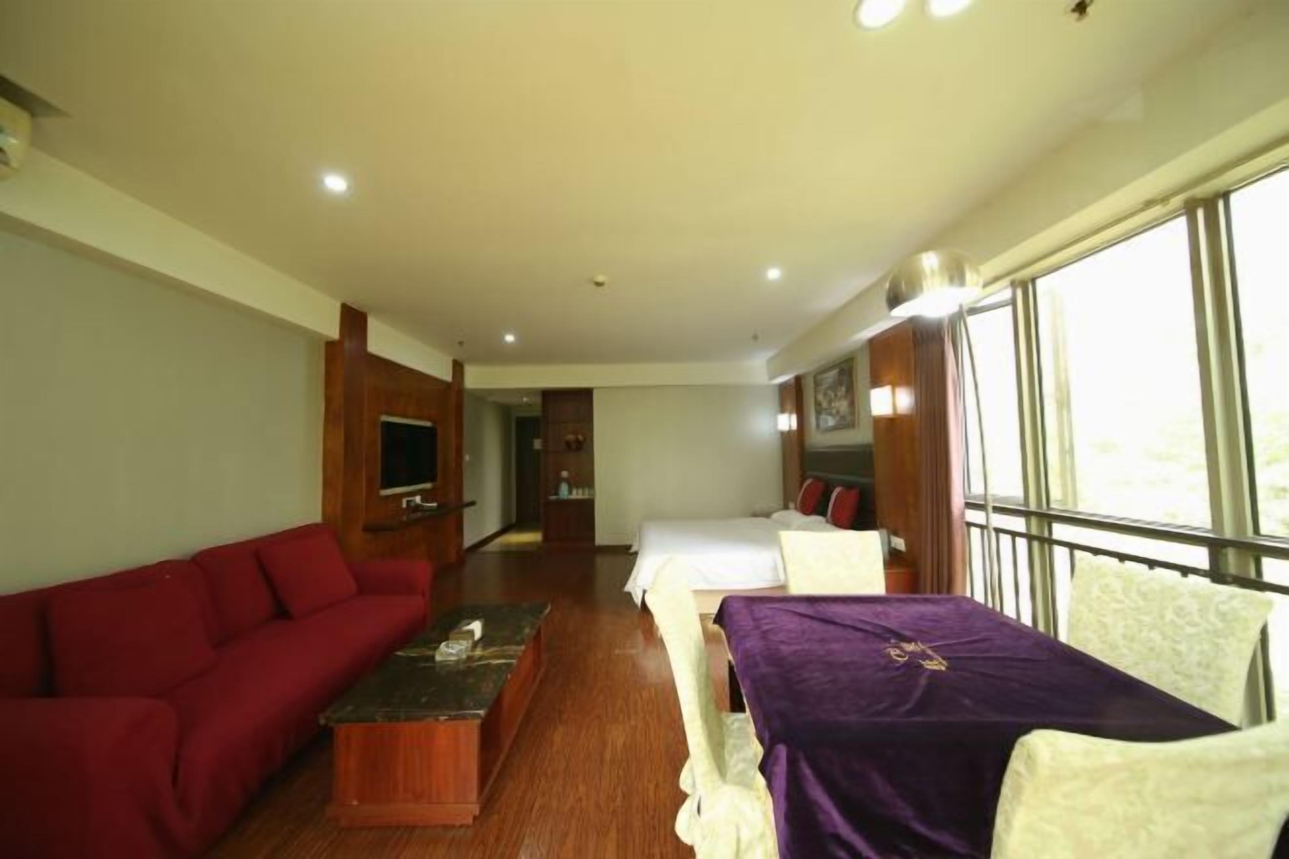 Habitación de hotel con un sofá rojo, una mesa de centro de madera, una mesa morada con una silla blanca, una cama con ropa de cama blanca y un ventanal con vistas.