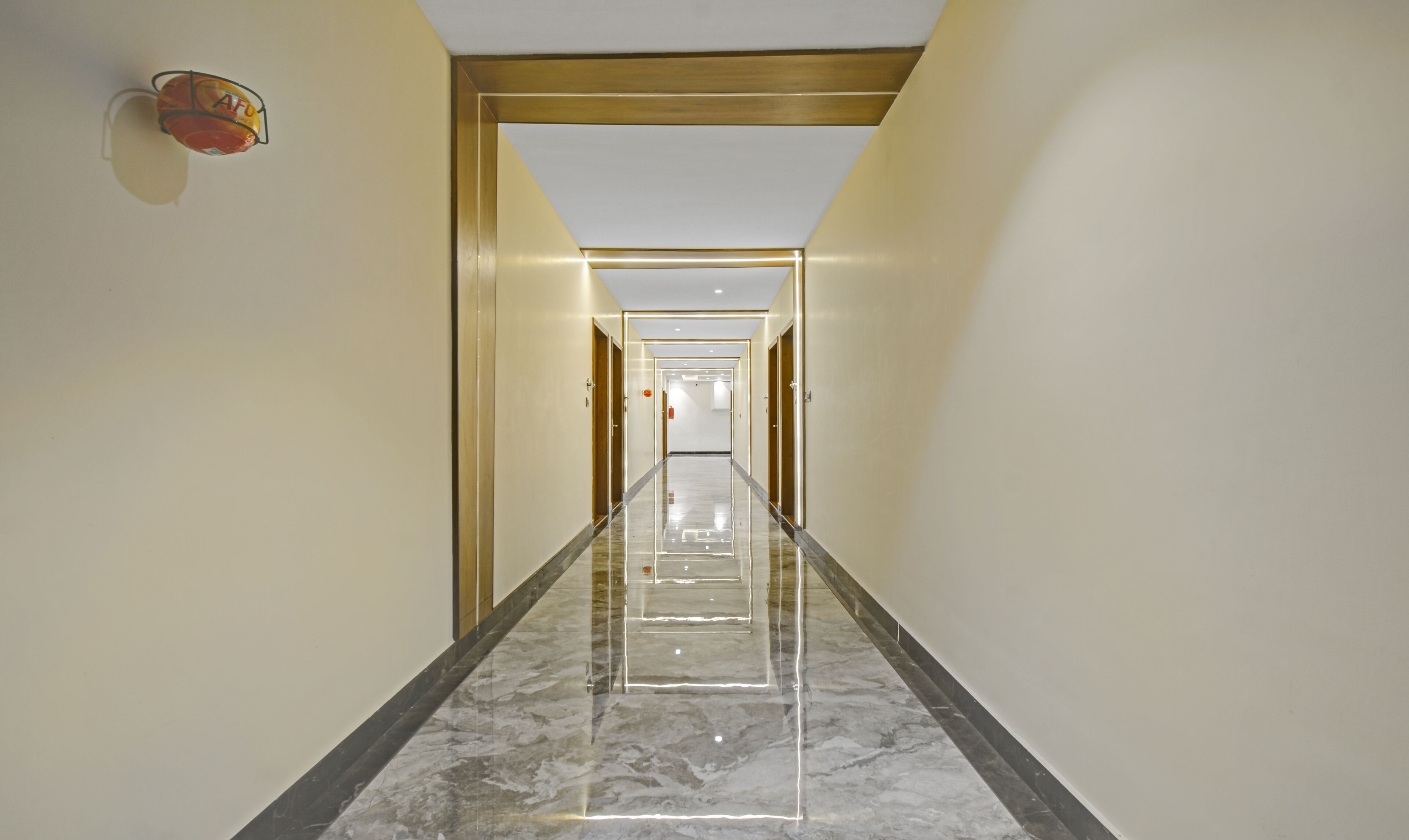 A long, narrow hallway with marble flooring and gold-framed doors, leading to a bright room with a white ceiling and recessed lighting.