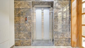 Un ascensor con pantalla digital y puerta de vidrio transparente, situado en un pasillo de paredes de mármol.