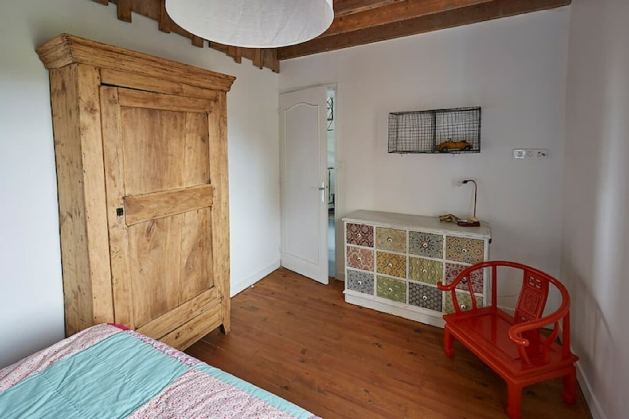 Una habitación con un armario de madera, un mueble de almacenaje colorido, una silla roja y una cama con colcha.