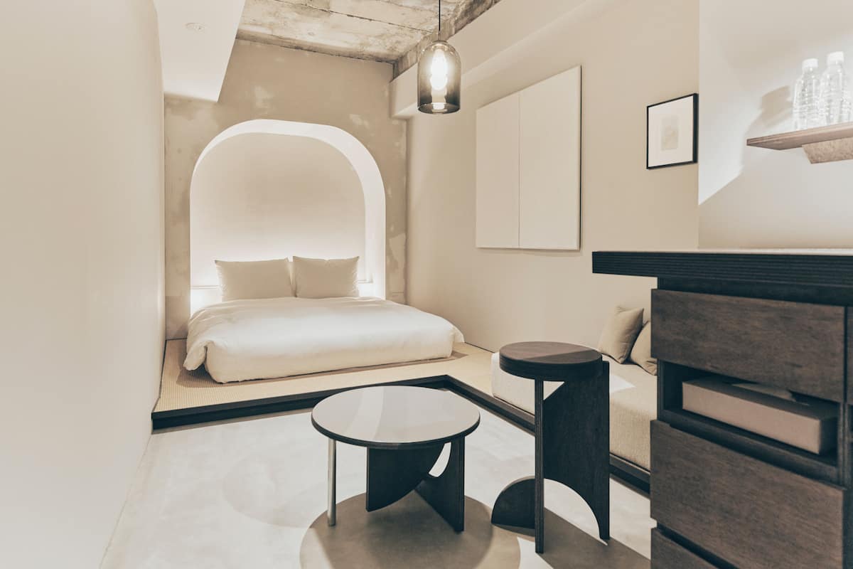 Una habitación de hotel moderna con una cama, una mesa redonda, una mesita auxiliar pequeña, un aparador de madera y un candelabro colgante.