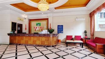 Vestíbulo de hotel con mostrador de recepción, área de check-in, zona de estar con sofá y sillones, y piso estampado.