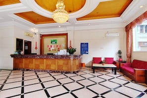 Um hall de hotel com balcão de recepção, mesa de check-in, área de estar com sofá e poltronas, e piso estampado.