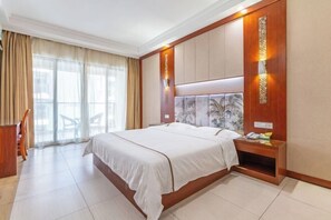 A hotel room with a large bed, a wooden nightstand, a desk, and a view of the outside through large windows. - Xingyu Sea View Hotel (Sanya Nanbian, Hailu Huitou) (Sanya)