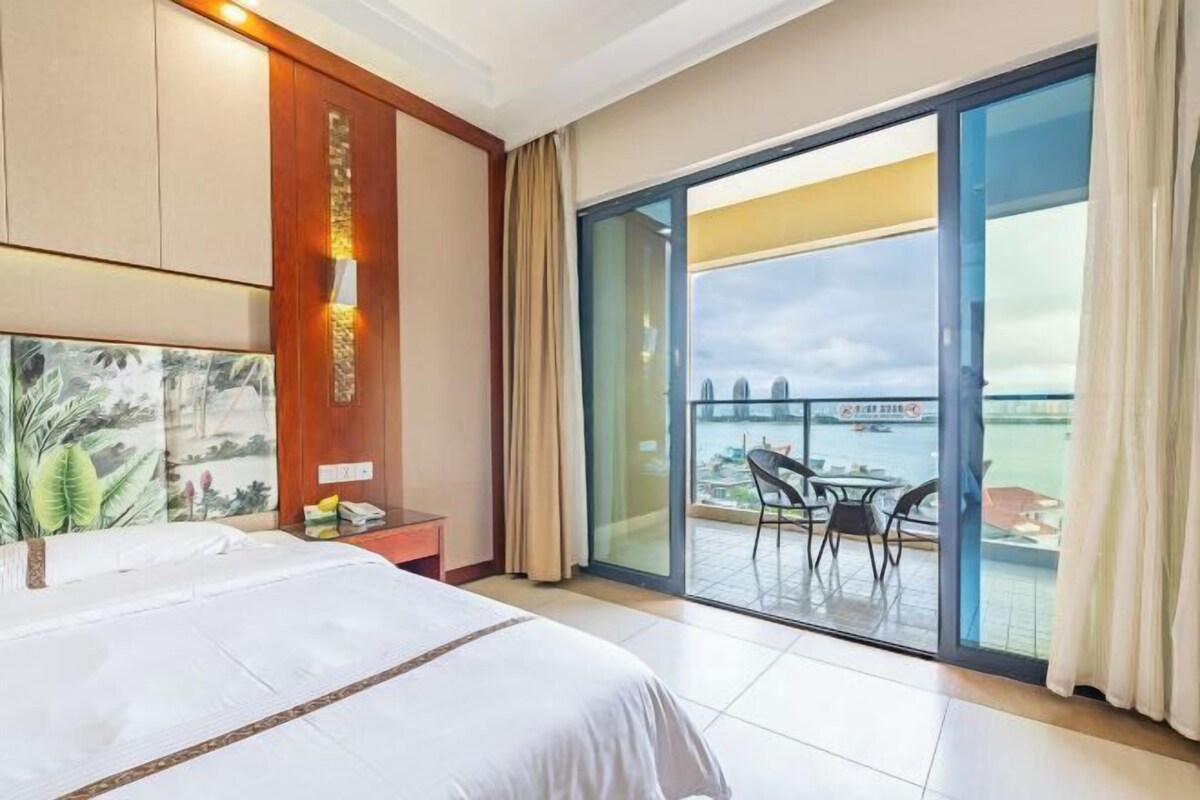 A hotel room with a large bed, a nightstand, a wall-mounted lamp, and a balcony with a table and chairs.