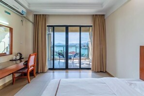 A hotel room with a bed, a desk with a chair, a large window with curtains, and a view of the sea and mountains. - Xingyu Sea View Hotel (Sanya Nanbian, Hailu Huitou) (Sanya)