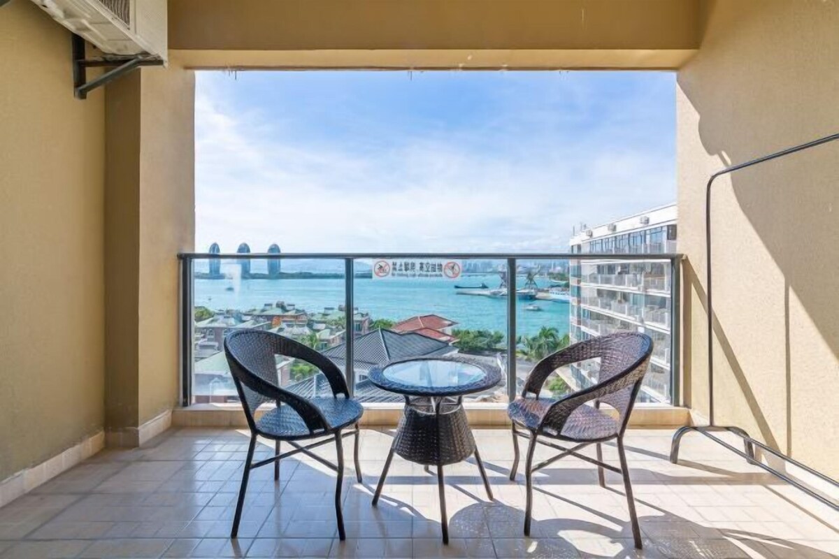 A balcony with two wicker chairs and a small round table, overlooking a coastal view with a bridge and buildings.