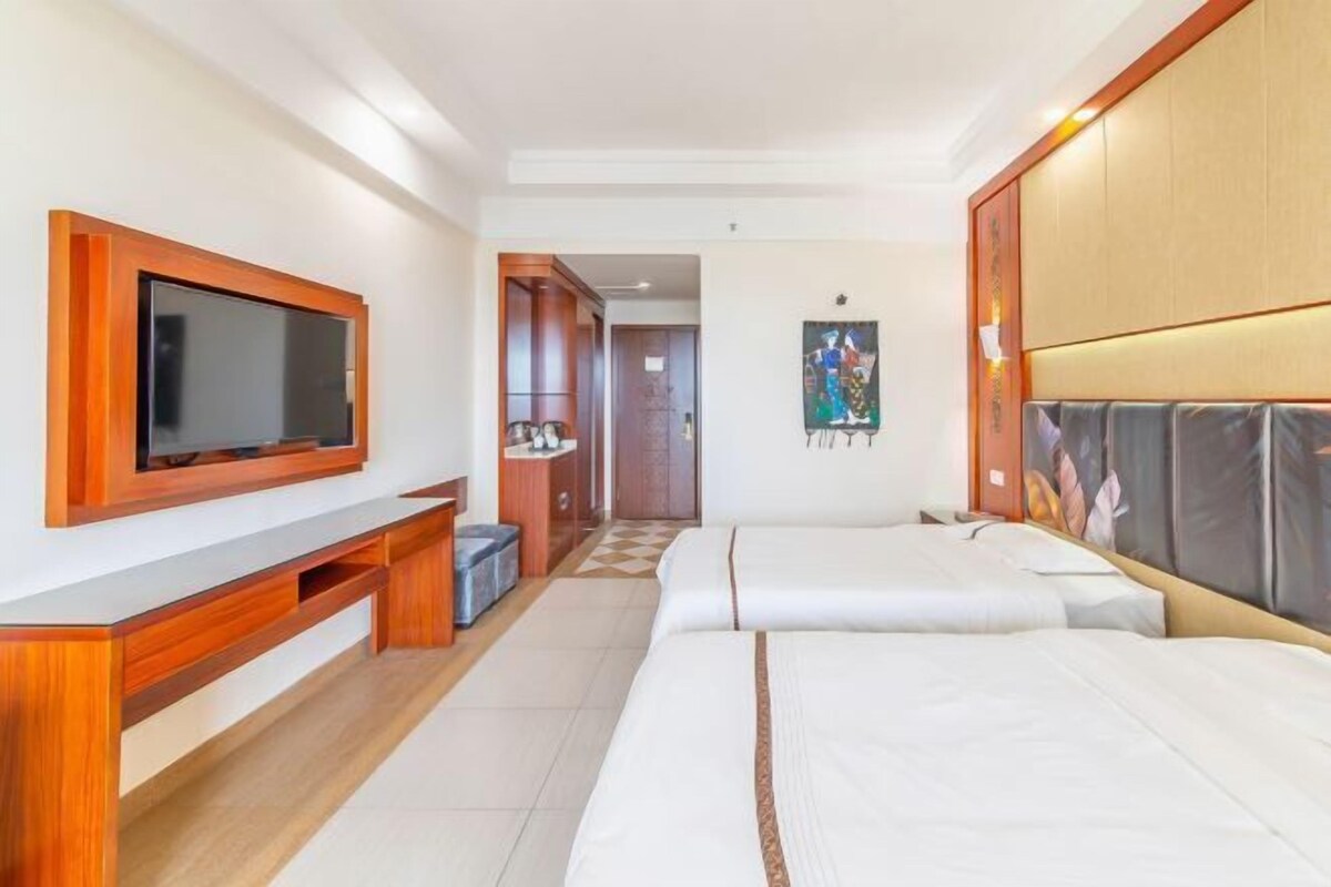 A hotel room with a large flat-screen TV mounted on the wall, a wooden desk, a bed with white bedding, and a headboard with a cushioned backrest.