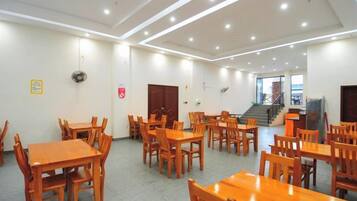 A spacious dining area with multiple wooden tables and chairs, a staircase in the background, and a fire alarm on the wall.