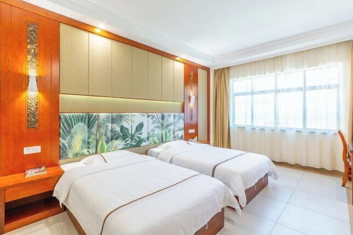 A hotel room with two beds, a large window with curtains, a wooden headboard, and a wall-mounted lamp.