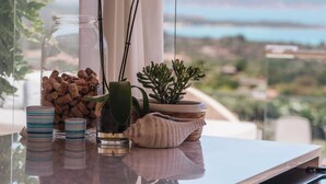 Habitació amb vista al mar, una làmpada de vímet, orquídies blanques, un brolla de vidre amb taps i una petita planta en test sobre una superfície reflectant.