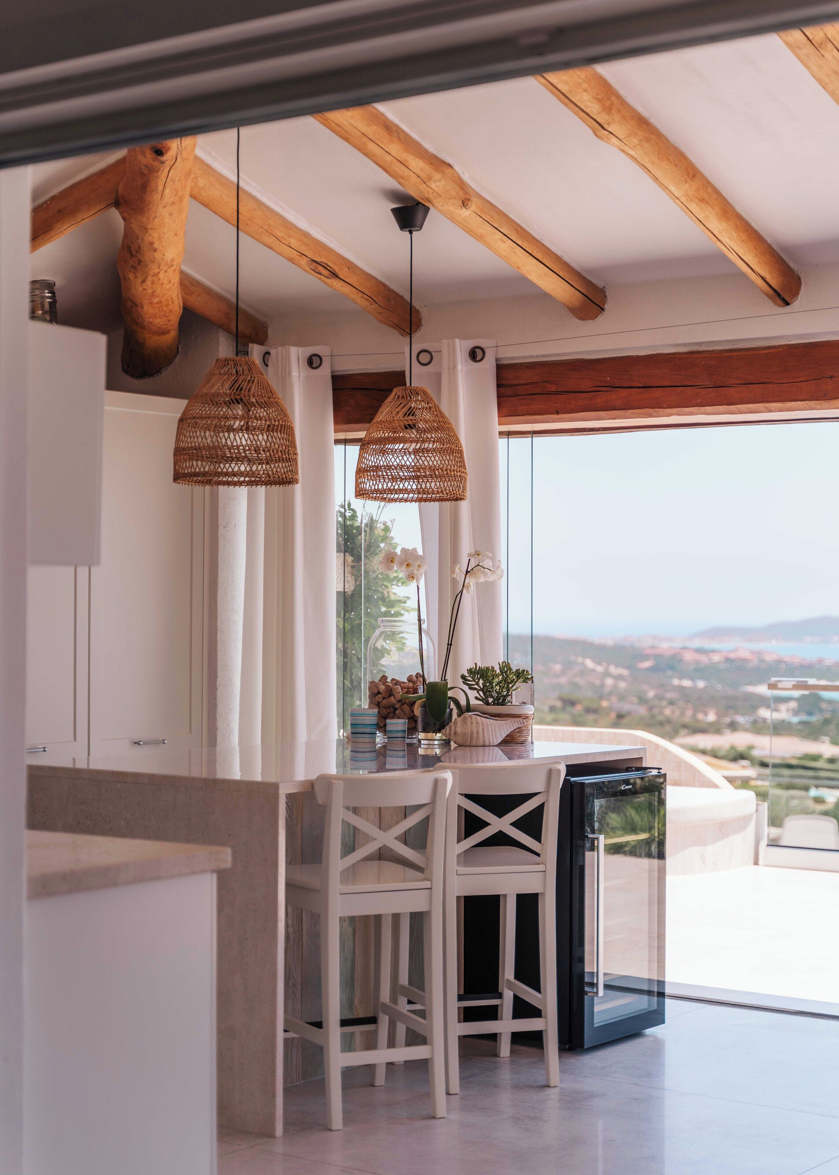 A modern kitchen with a large window, wooden beams, and two wicker pendant lights.