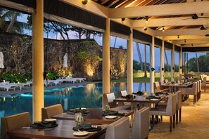 Un área de comedor al aire libre con mesas y sillas, una piscina y vistas a un paisaje rocoso con árboles.