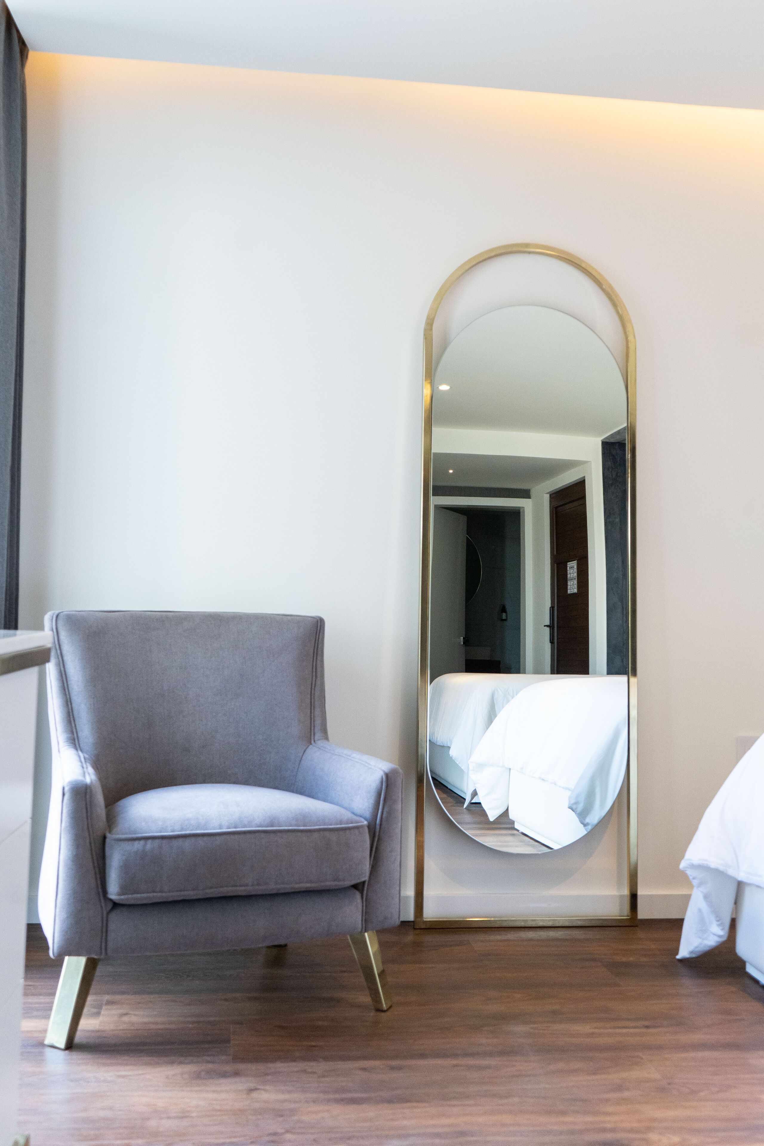 A modern room with a grey armchair, a large mirror, and a bed with white bedding.