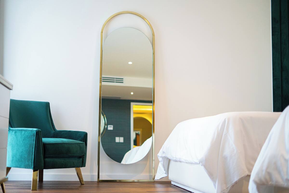 Une chambre d’hôtel avec un fauteuil turquoise, un grand miroir et un lit recouvert de linge de lit blanc.