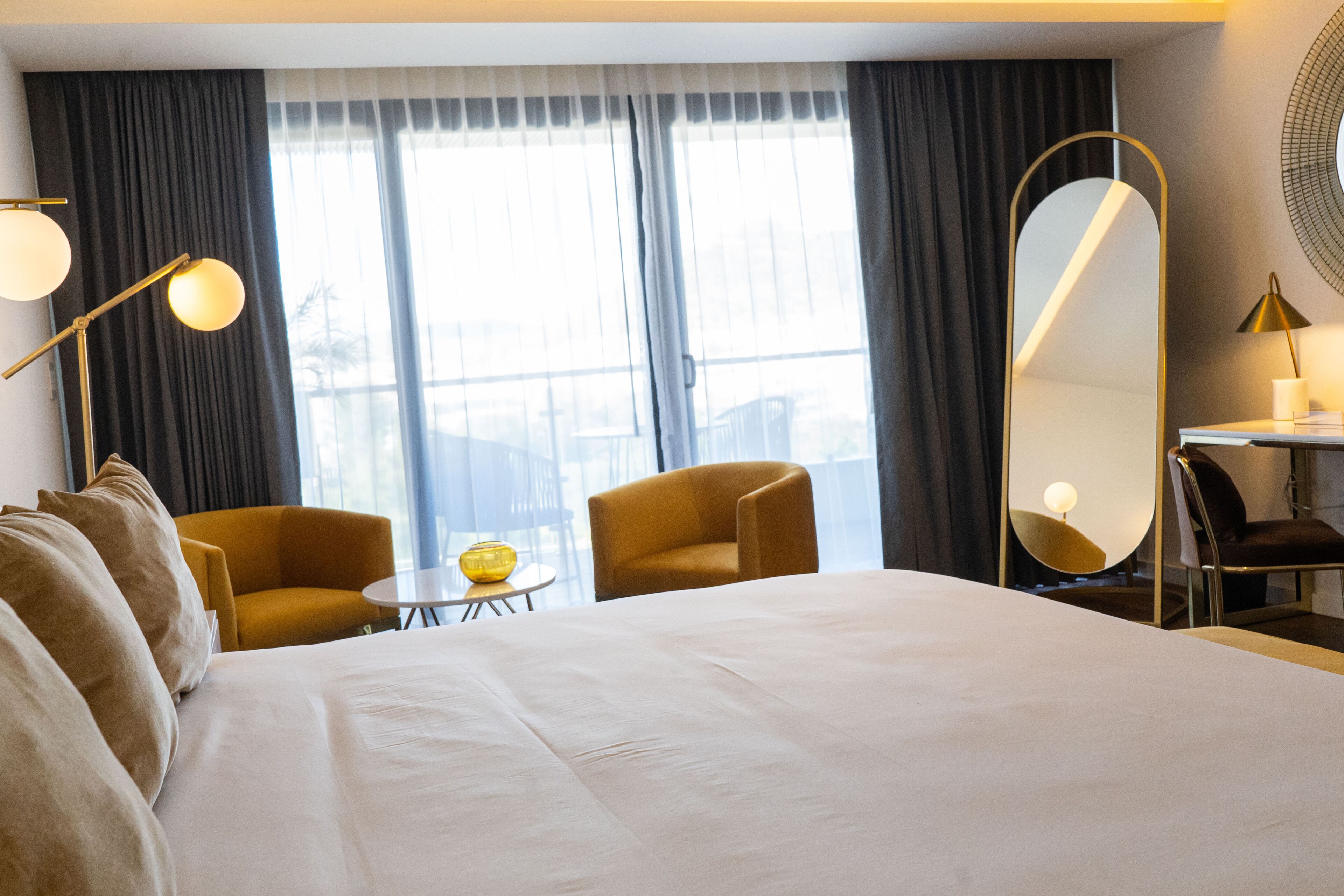 Habitación de hotel con una cama grande, dos sillones, una mesita, un espejo y una ventana con cortinas.