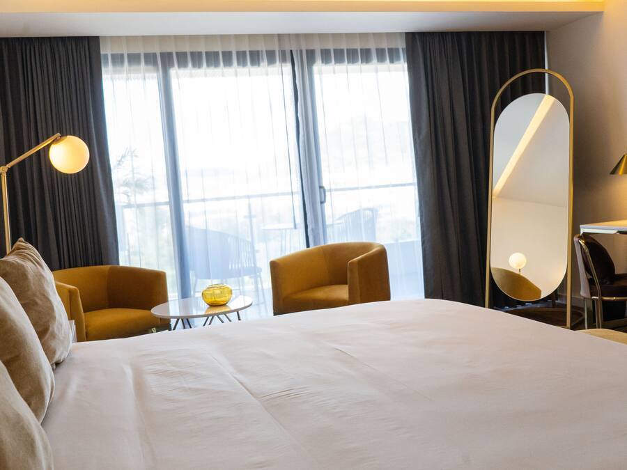 A hotel room with a large bed, two armchairs, a small table, a mirror, and a window with curtains.