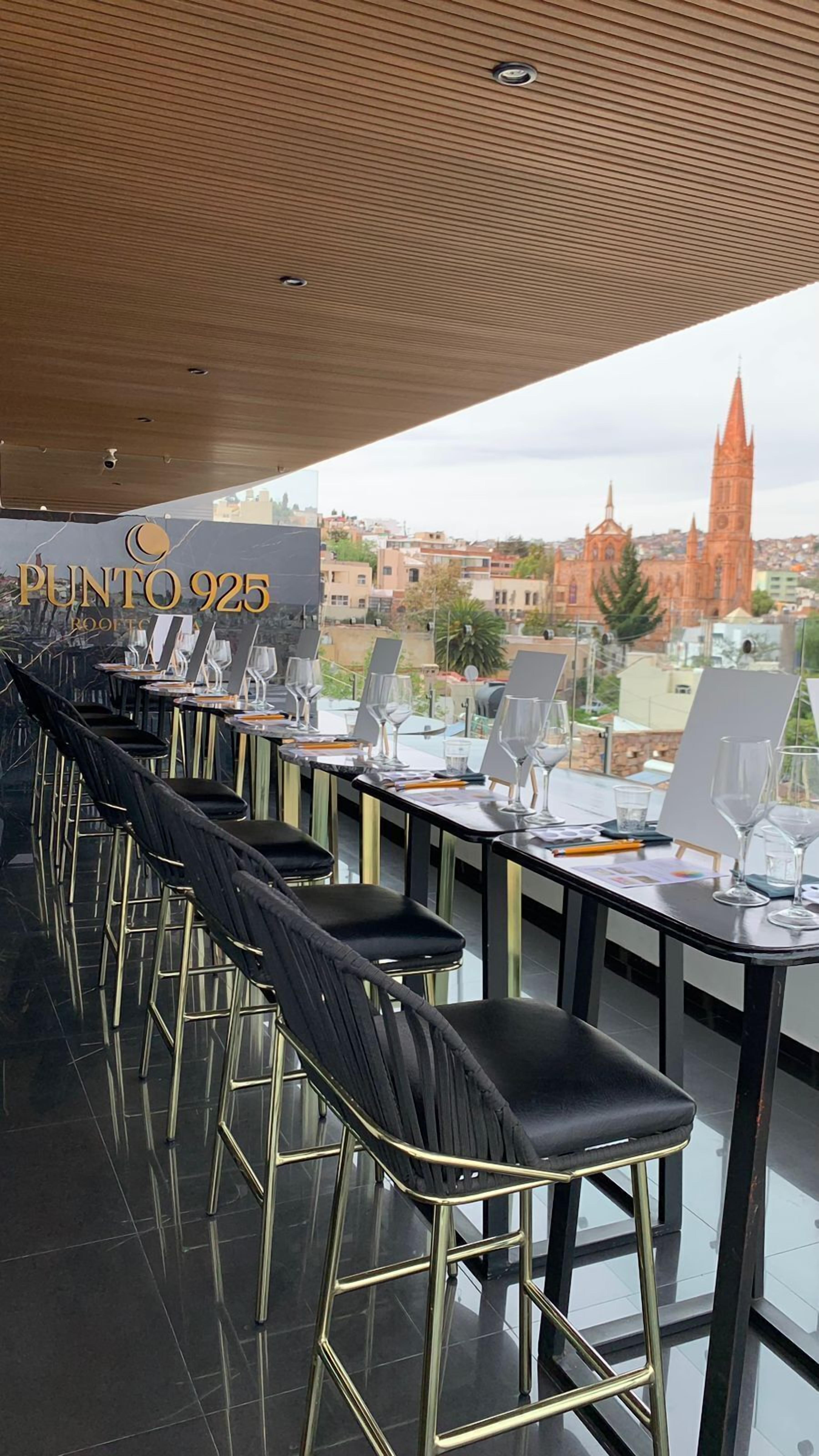 A rooftop bar with black leather chairs and tables, a view of a cityscape including a church tower, and the text "PUNTO 925 ROOFTOP".