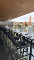 Un bar en la azotea con sillas y mesas de cuero negro, una vista de la ciudad que incluye una torre de iglesia, y el texto "PUNTO 925 ROOFTOP".