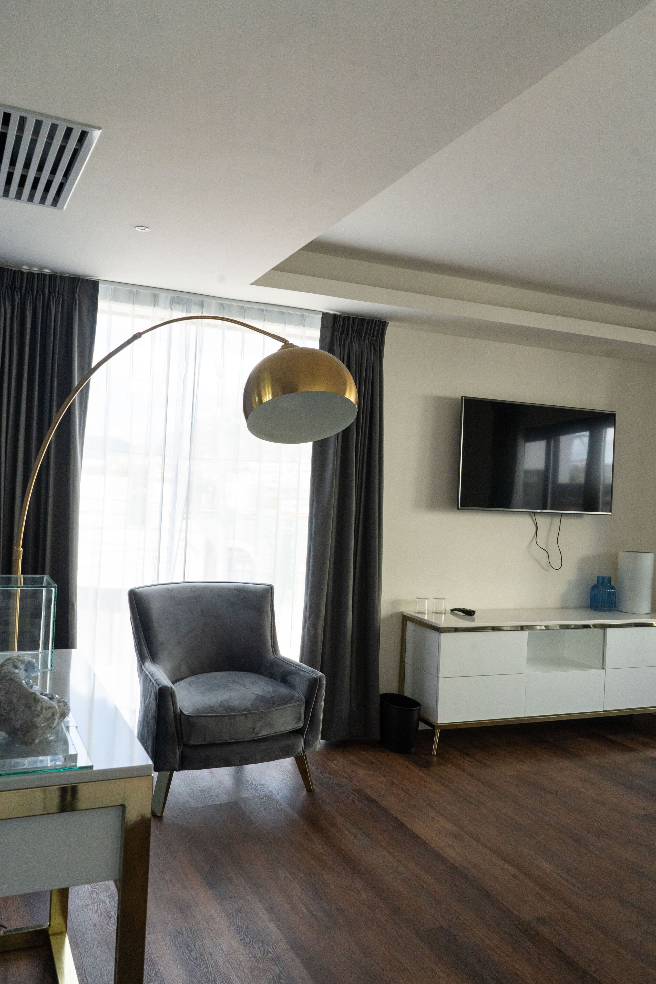A modern living room with a dark grey armchair, a white console table, a glass coffee table, a floor lamp, a flat-screen TV mounted on the wall, and a large window with curtains.