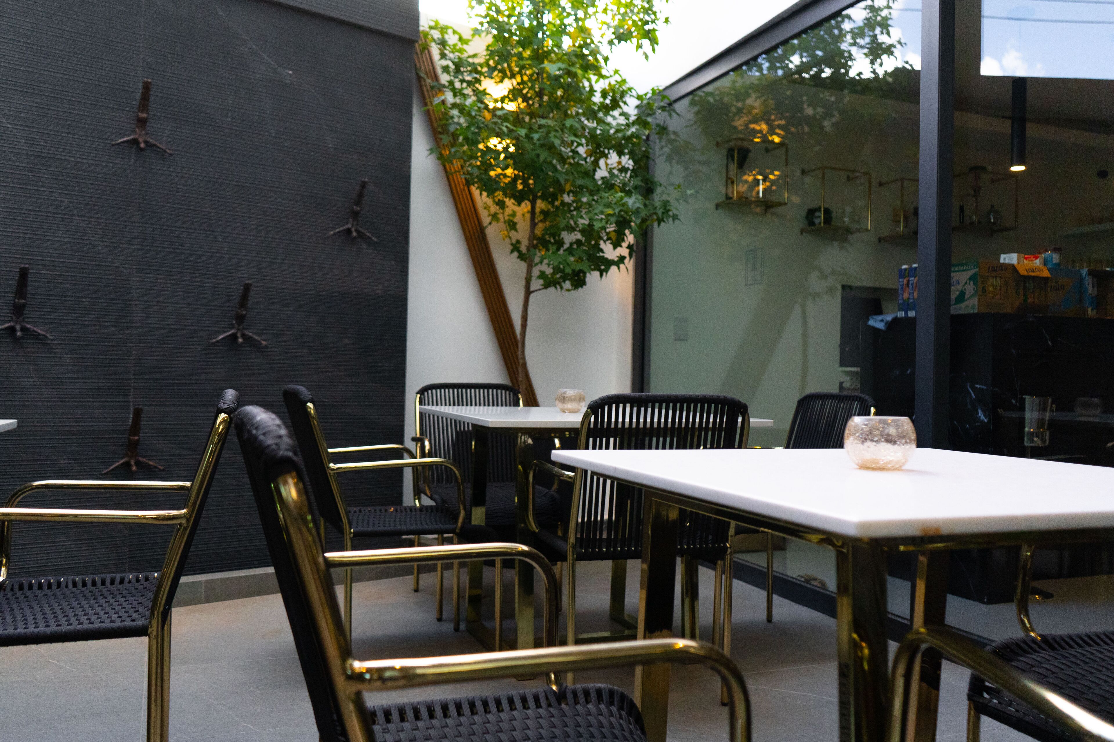 An outdoor seating area with tables and chairs, a tree, and a glass wall reflecting the interior.