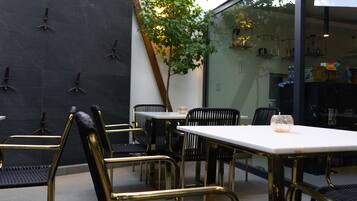 An outdoor seating area with tables and chairs, a tree, and a glass wall reflecting the interior.
