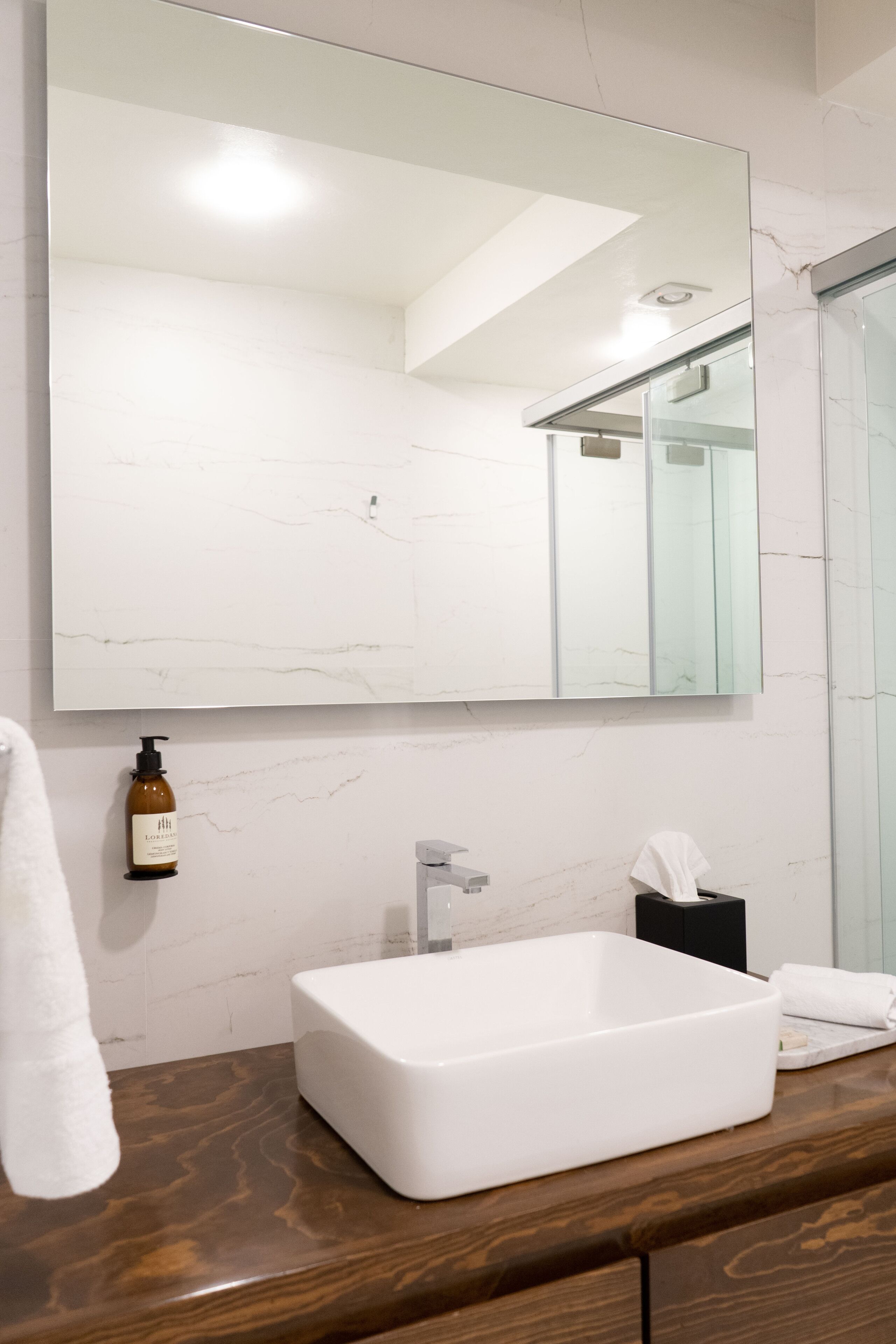 A bathroom with a white marble wall, a large mirror, a white sink, and a wooden countertop.