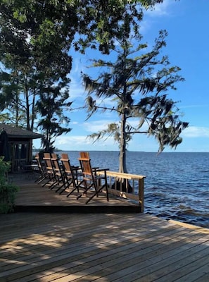 Une terrasse en bois avec des chaises donnant sur une étendue d’eau, le tout entouré d’arbres.