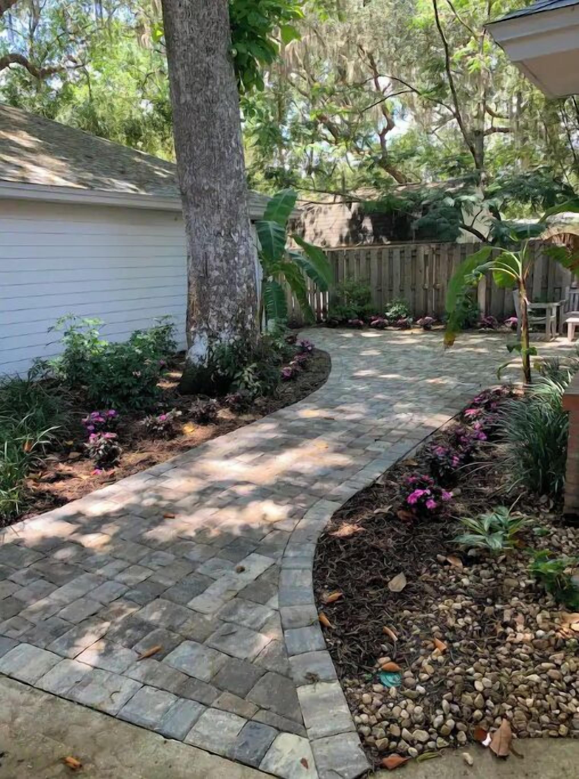 A paved pathway leading to a house with a wooden fence and a tree.