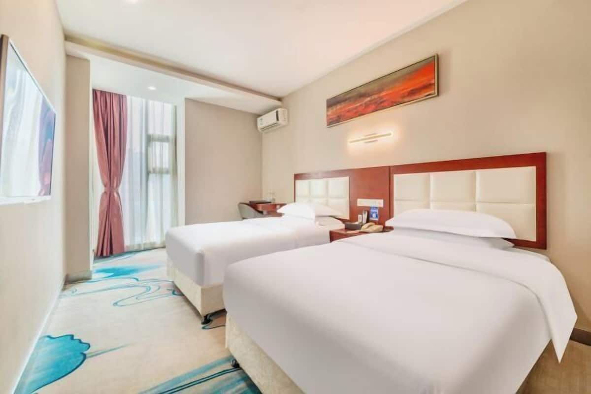 A hotel room with two beds, a large window with curtains, a painting on the wall, and a carpet with a blue pattern.