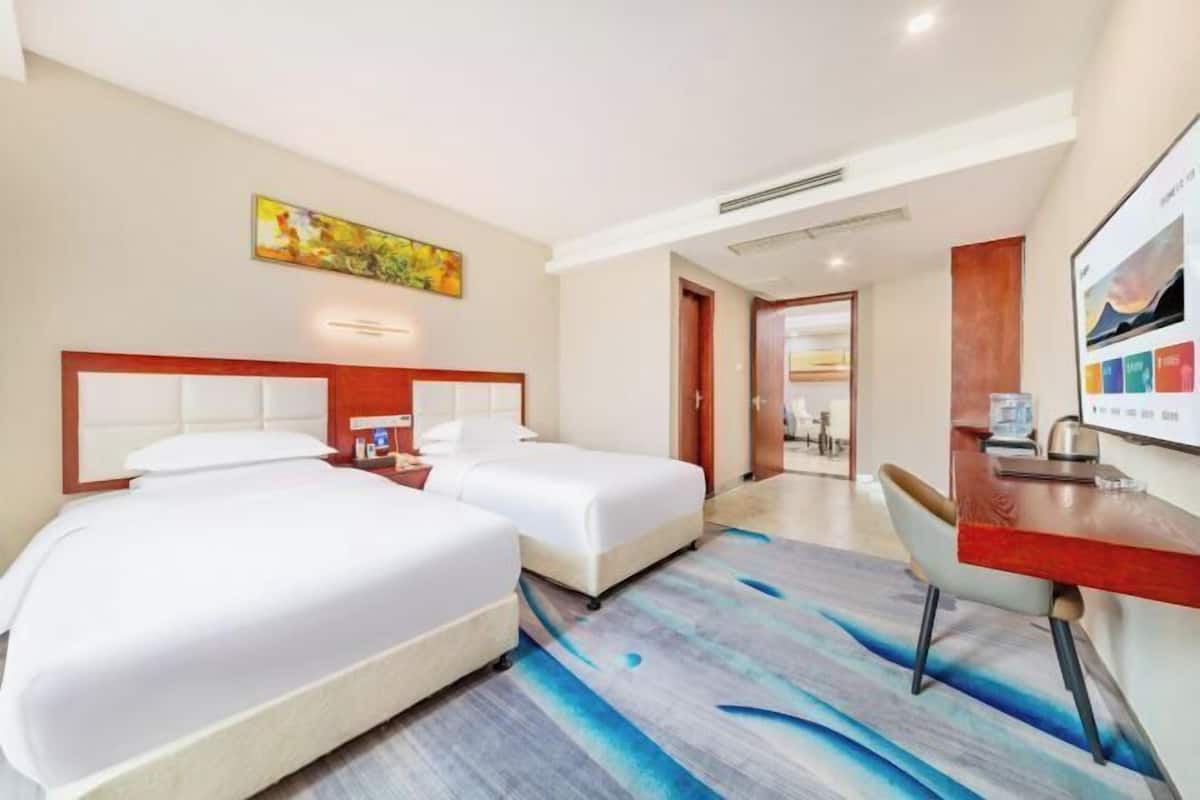 A hotel room with two beds, a desk, and a chair. There is a painting on the wall and a window with a view.