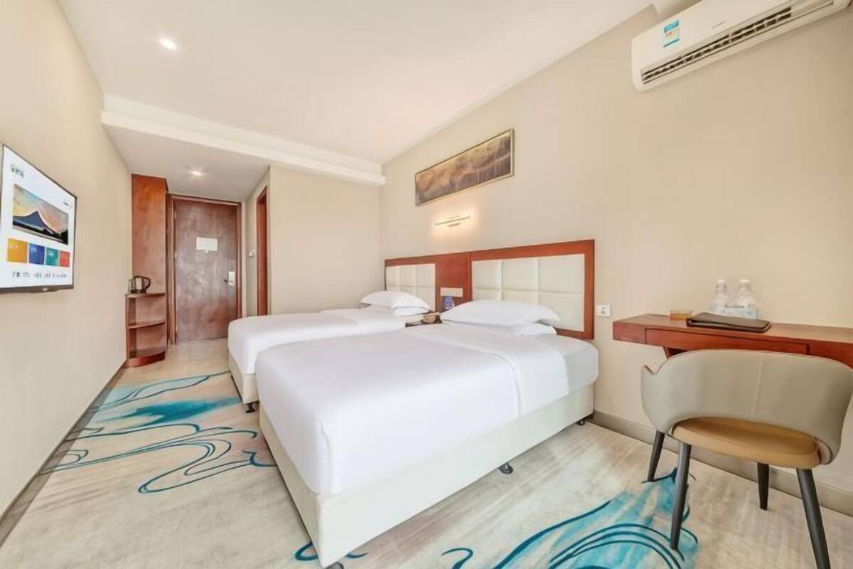 A hotel room with two beds, a desk, a chair, a painting, and an air conditioning unit.