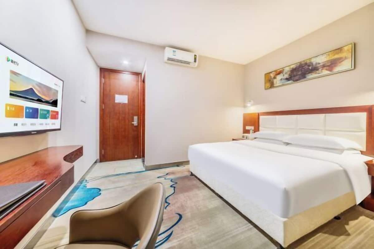 A hotel room with a bed, a desk, a chair, an air conditioning unit, and a TV displaying a colorful interface.