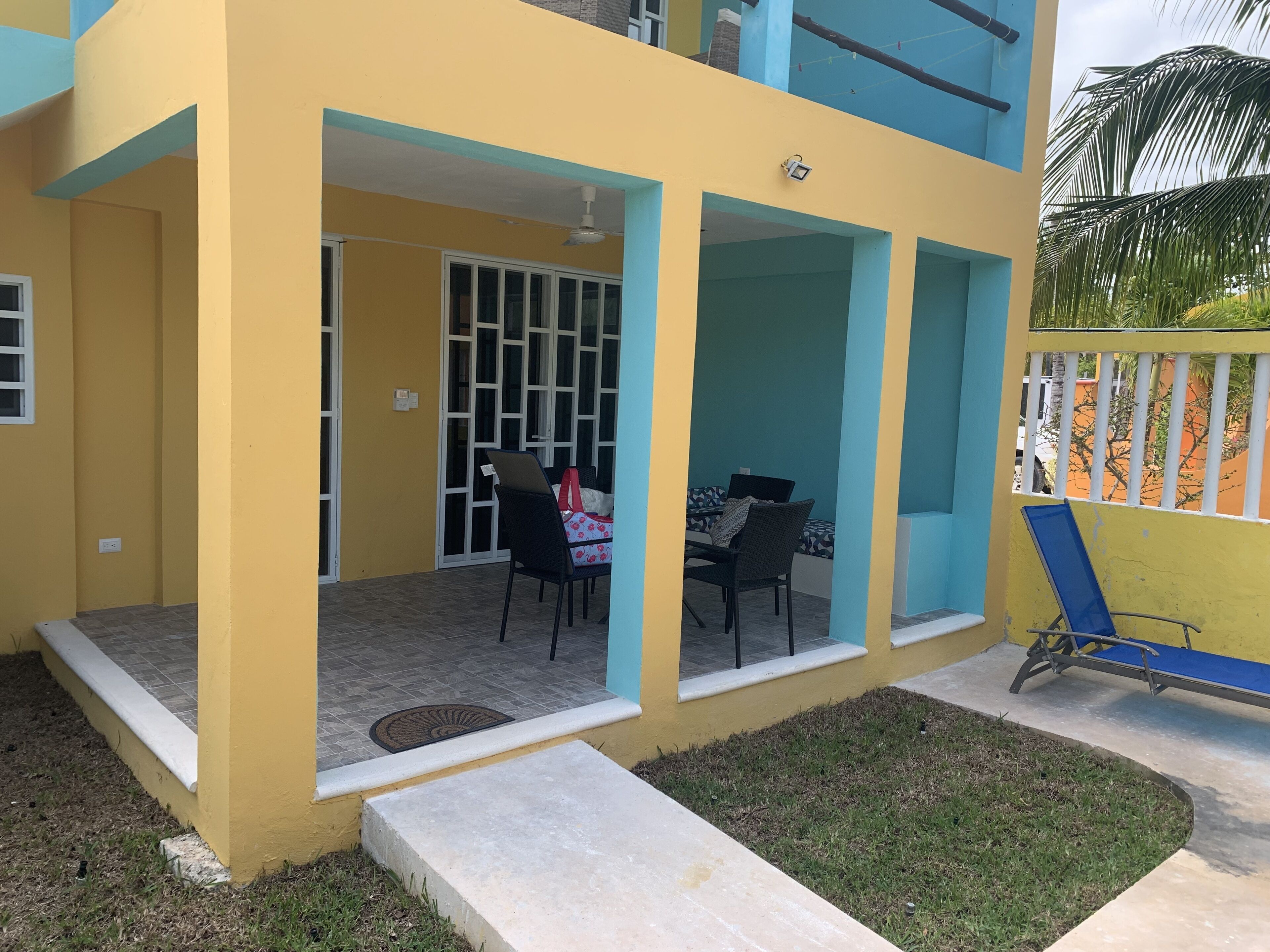 A two-story building with a yellow exterior and blue accents. There is a covered patio area with a table and chairs, and a blue chair leaning against the wall.